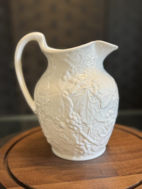 A white ceramic pitcher with an embossed grapevine and leaf pattern sits on a round wooden surface. The pitcher has a curved handle, a wide spout, and a glossy finish. The background is softly blurred and dark, making the pitcher stand out.