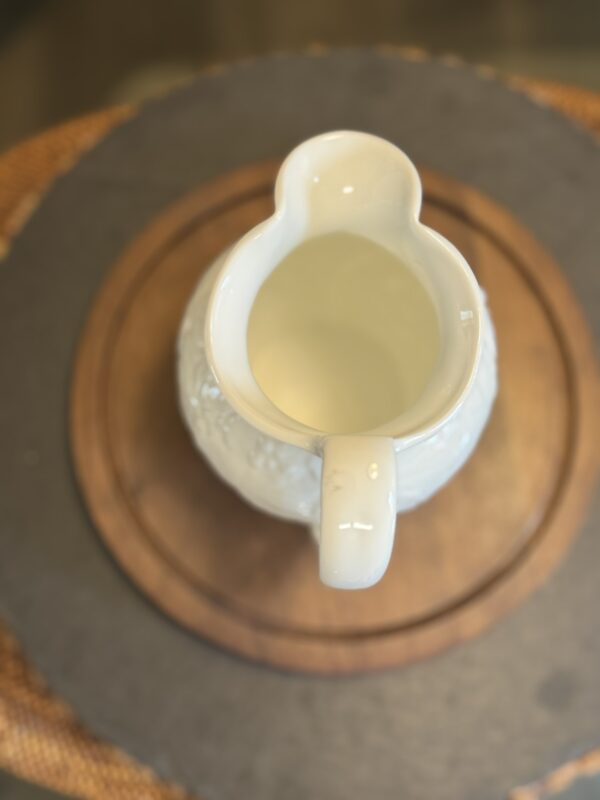 A white ceramic pitcher with a textured design sits on top of two stacked round wooden trays, viewed from above. The background is softly blurred, drawing focus to the pitcher as the central subject.