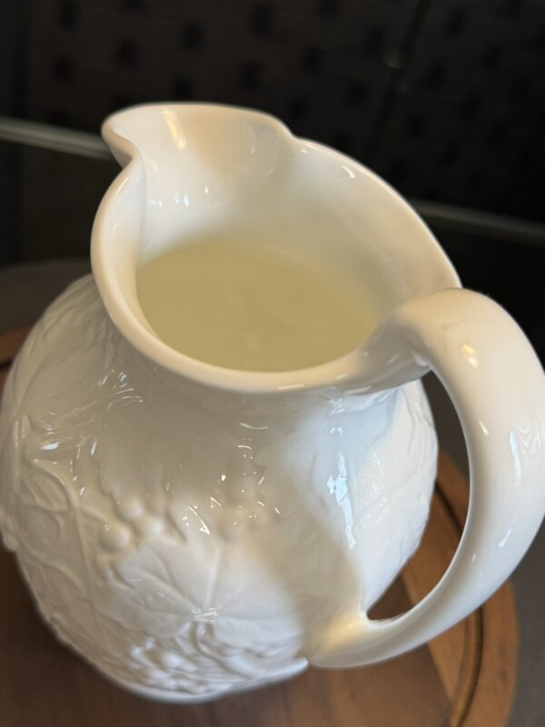 A close-up of a white ceramic pitcher with embossed floral patterns, filled with a pale, translucent liquid. The pitcher sits on a round wooden tray, and the background is softly blurred, creating a gentle, elegant atmosphere.
