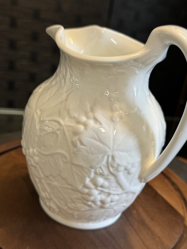 A white ceramic pitcher with a glossy finish sits on a wooden surface. The pitcher features embossed grapevine and leaf patterns on its surface, a curved spout, and a large, graceful handle. The background is softly blurred and dark-toned.