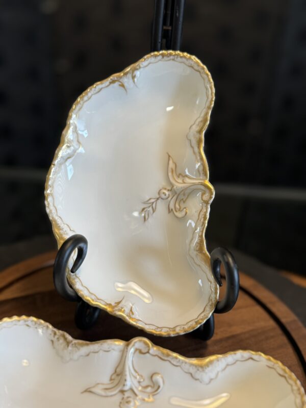 An elegant, irregularly shaped porcelain dish with a white glaze, gold trim, and ornate gold floral pattern, displayed upright on a black stand. The background is softly blurred, highlighting the dishs details.