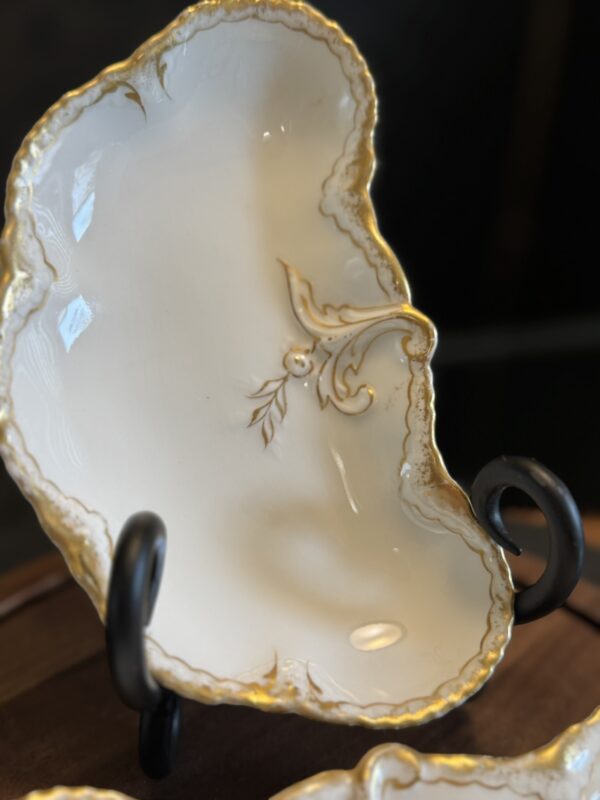 An ornate, cream-colored porcelain dish with gold trim and decorative gold detailing rests on a black stand. The dish has an irregular, organic shape with a wavy rim and embossed patterns near the edge, displayed against a dark, blurred background.