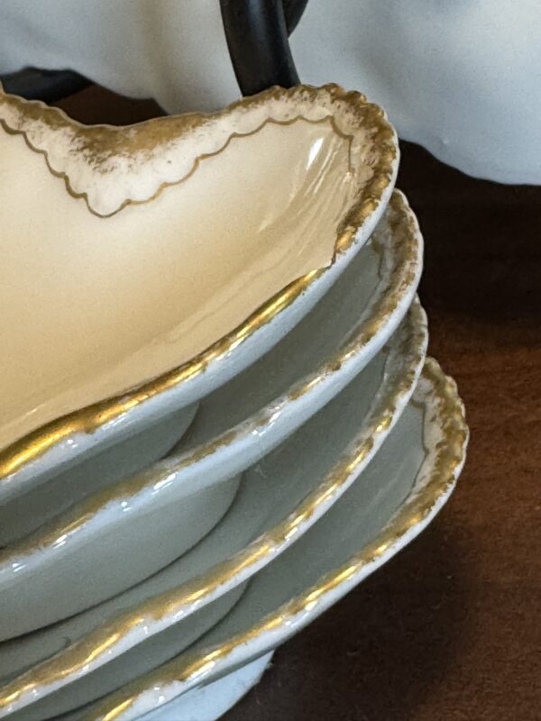 A close-up of a stack of four white porcelain dishes with scalloped, gold-trimmed edges. The dishes are arranged neatly on a brown surface, highlighting their elegant, vintage design and delicate details.