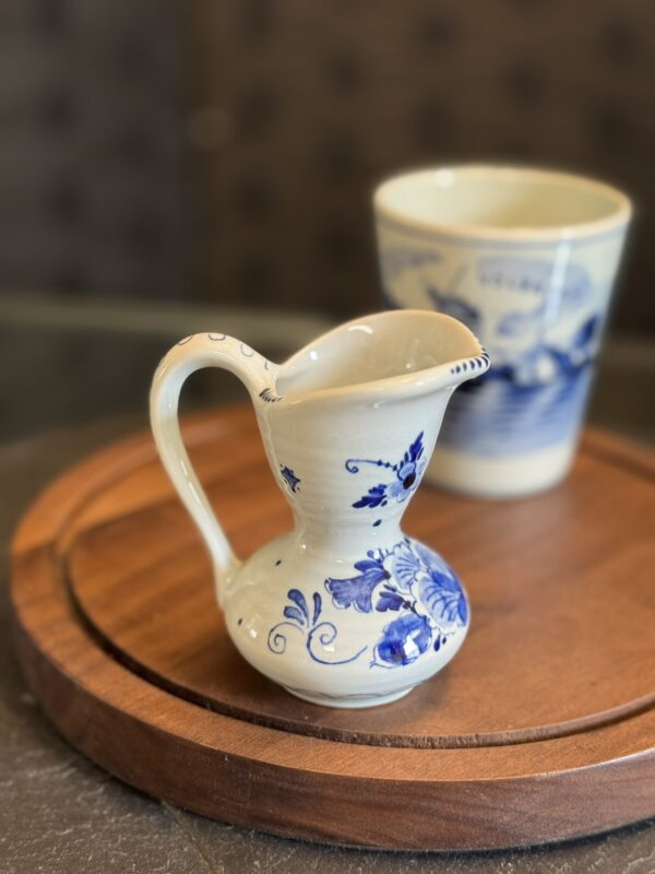 A small ceramic pitcher with a curved handle and blue floral patterns sits on a round wooden tray. Behind it, there is a matching cup decorated with a blue scenic design. The background is softly blurred and brown-toned.