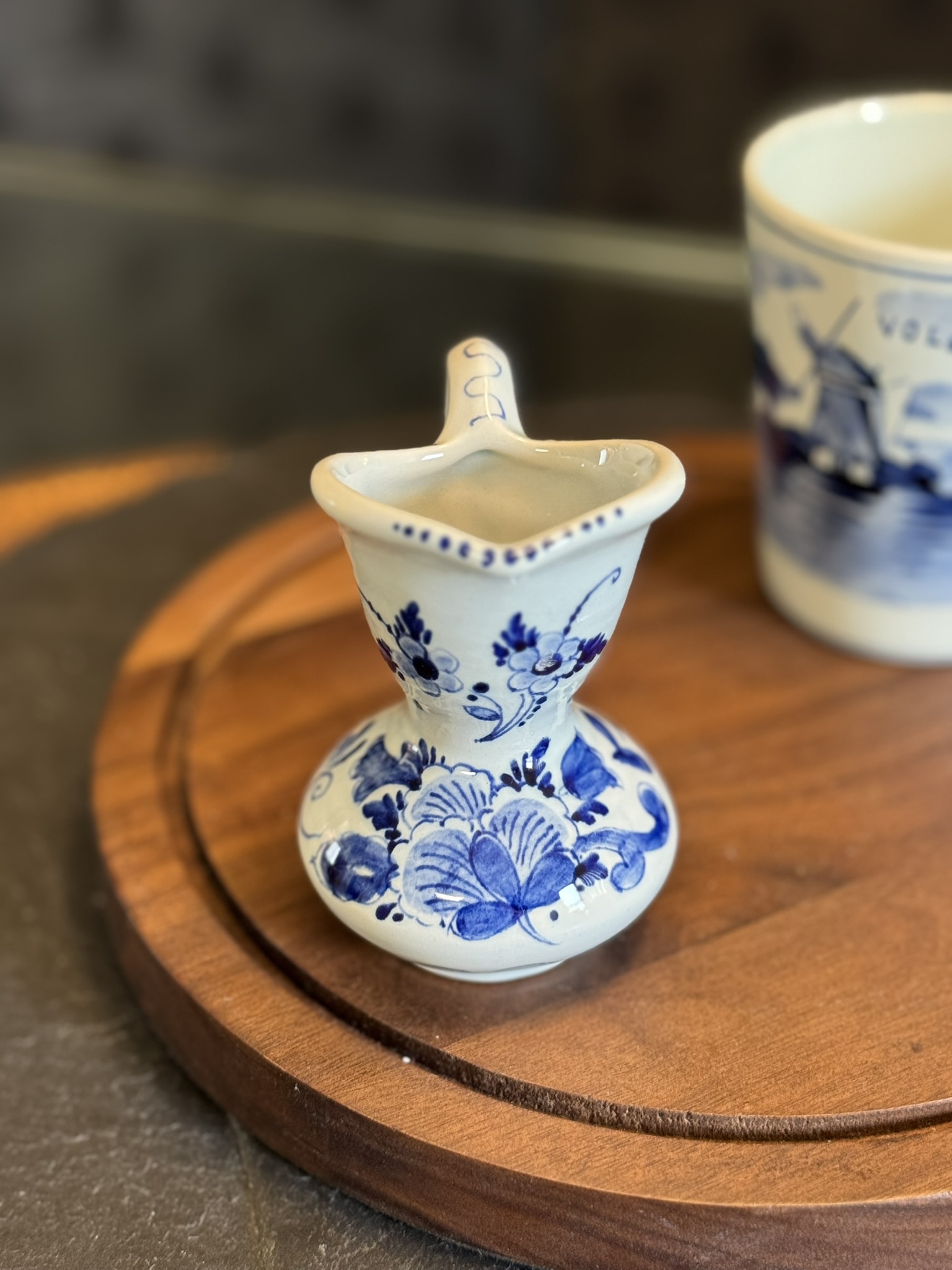 A small, ornately decorated blue and white ceramic pitcher with floral designs sits on a round wooden tray. The pitcher has a wide base, a narrow neck, a curved handle, and a spout. A cup with a blue scenic design is slightly blurred in the background.