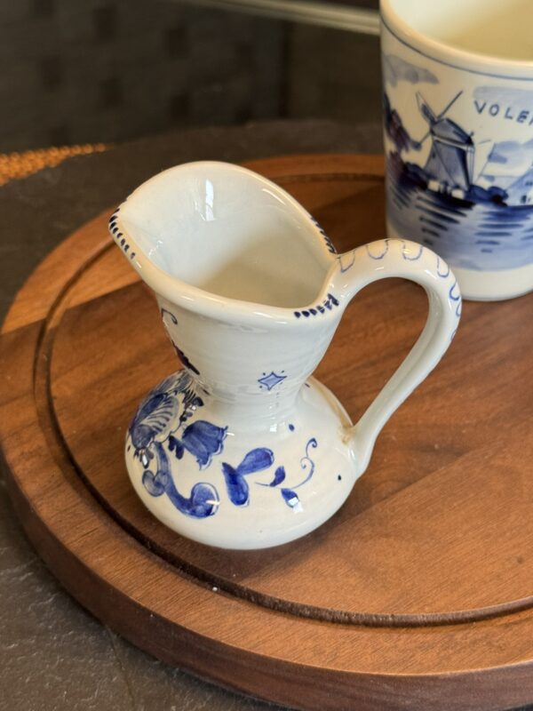 A small, white ceramic pitcher with blue floral patterns sits on a round wooden tray. Behind it is a white cup decorated with blue windmills and the word “VOLENDAM.” The background is blurred, highlighting the pitcher and cup.