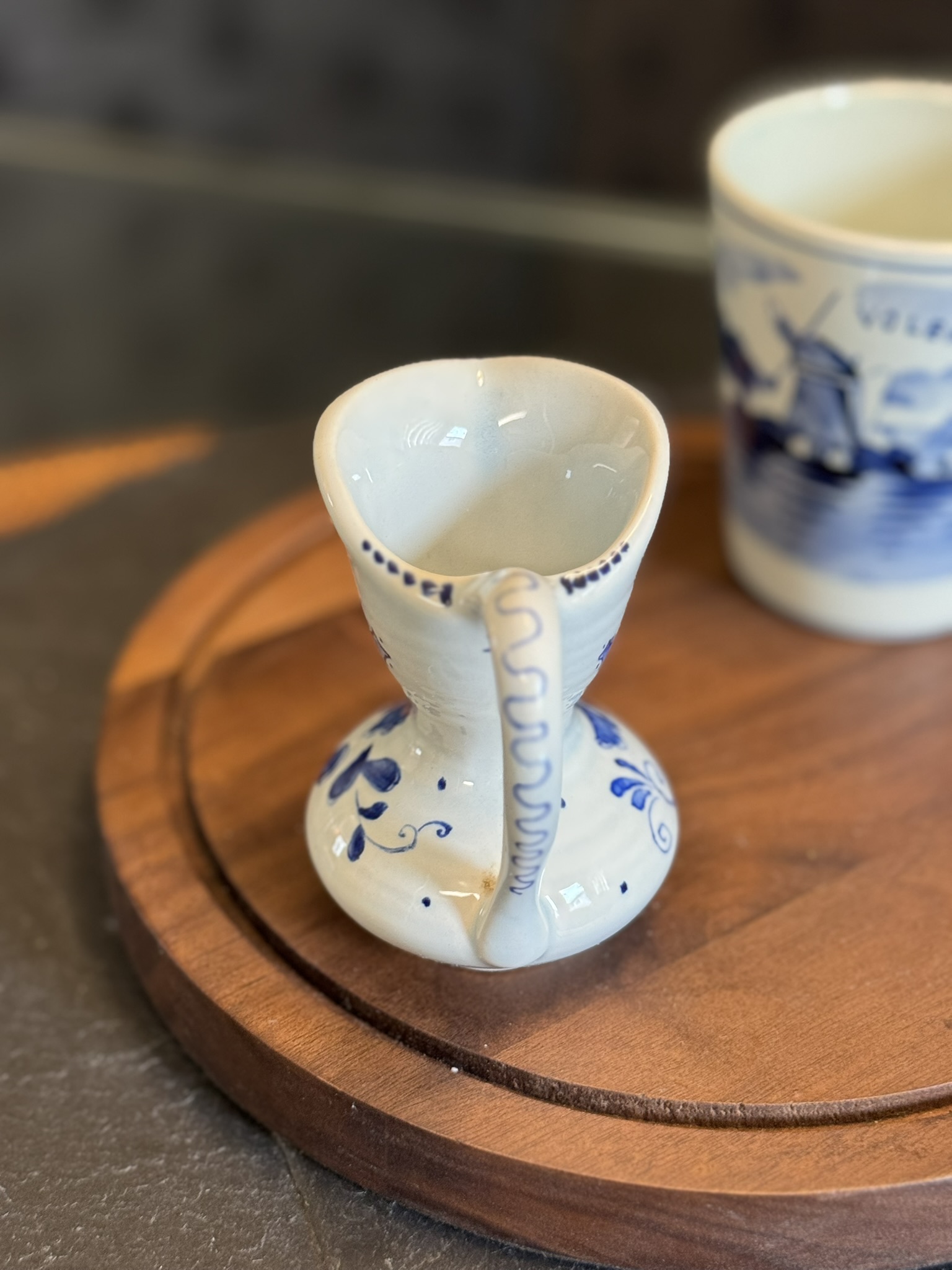 A small, white ceramic pitcher with blue floral designs and a delicate handle sits on a round wooden tray. In the blurred background, there is a larger cup with a blue nautical pattern. The surface beneath the tray appears to be dark stone.