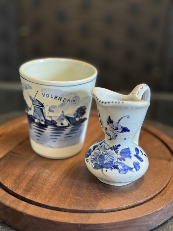 A small ceramic jug and a cup with blue Dutch-style designs sit on a round wooden tray. The cup features a windmill and the word Volendam; the jug is decorated with blue floral patterns. Both have a traditional handcrafted look.