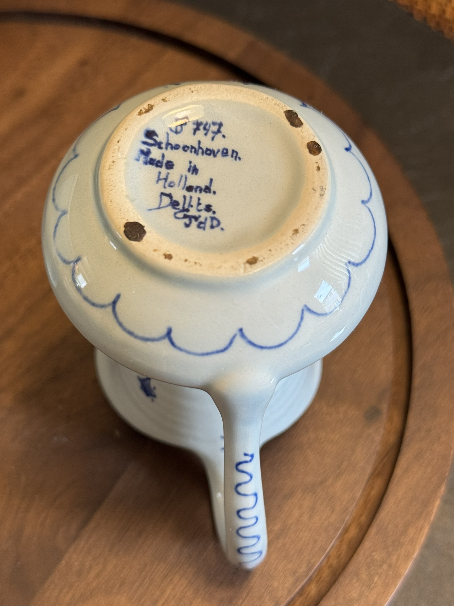 A small white ceramic cup with blue hand-painted designs sits upside down, revealing its base. Written in blue ink on the bottom are the words: “J847 Schoonhoven. Made in Holland. Delfts GY.D.” The cup rests on a round wooden surface.
