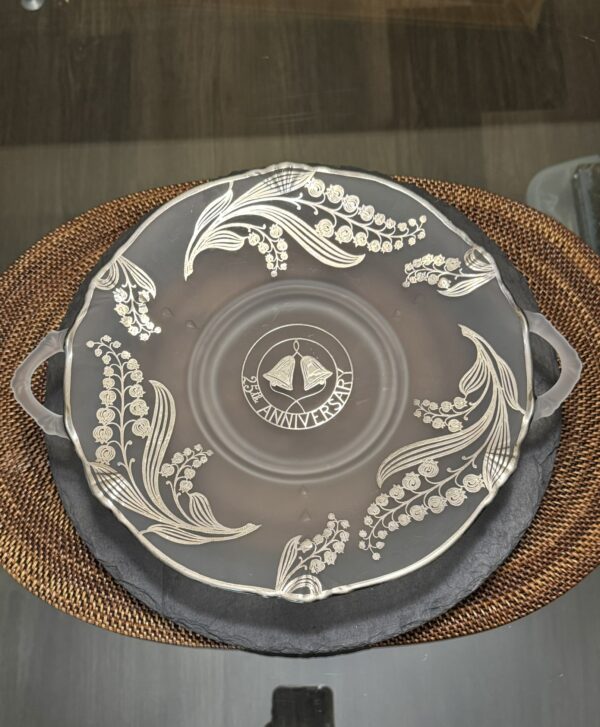 A round frosted glass platter with two small handles sits on a woven placemat. The platter features silver floral designs and a central emblem reading “25th Anniversary” with two bells and ribbon, symbolizing a silver wedding anniversary celebration.