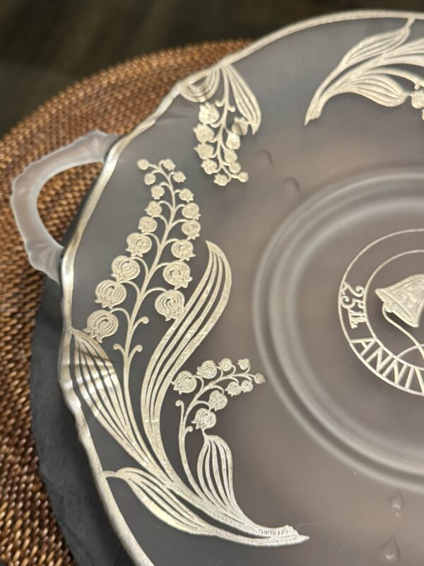 A frosted glass plate with an ornate silver floral design featuring lily of the valley flowers and leaves. The plate has a scalloped edge, a handle on the left, and a circular pattern with lettering at the center, resting on a textured brown surface.