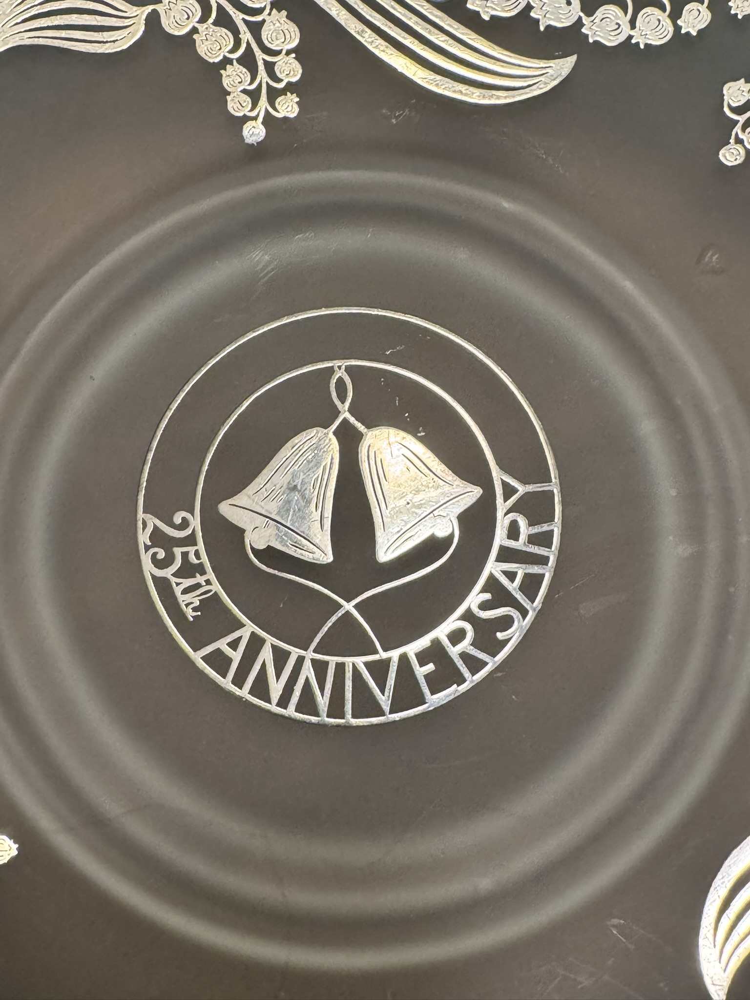 A decorative silver design on a dark surface features two bells inside a circle. The text reads 25th ANNIVERSARY curving around the bottom half of the circle. Floral patterns frame the top of the image.