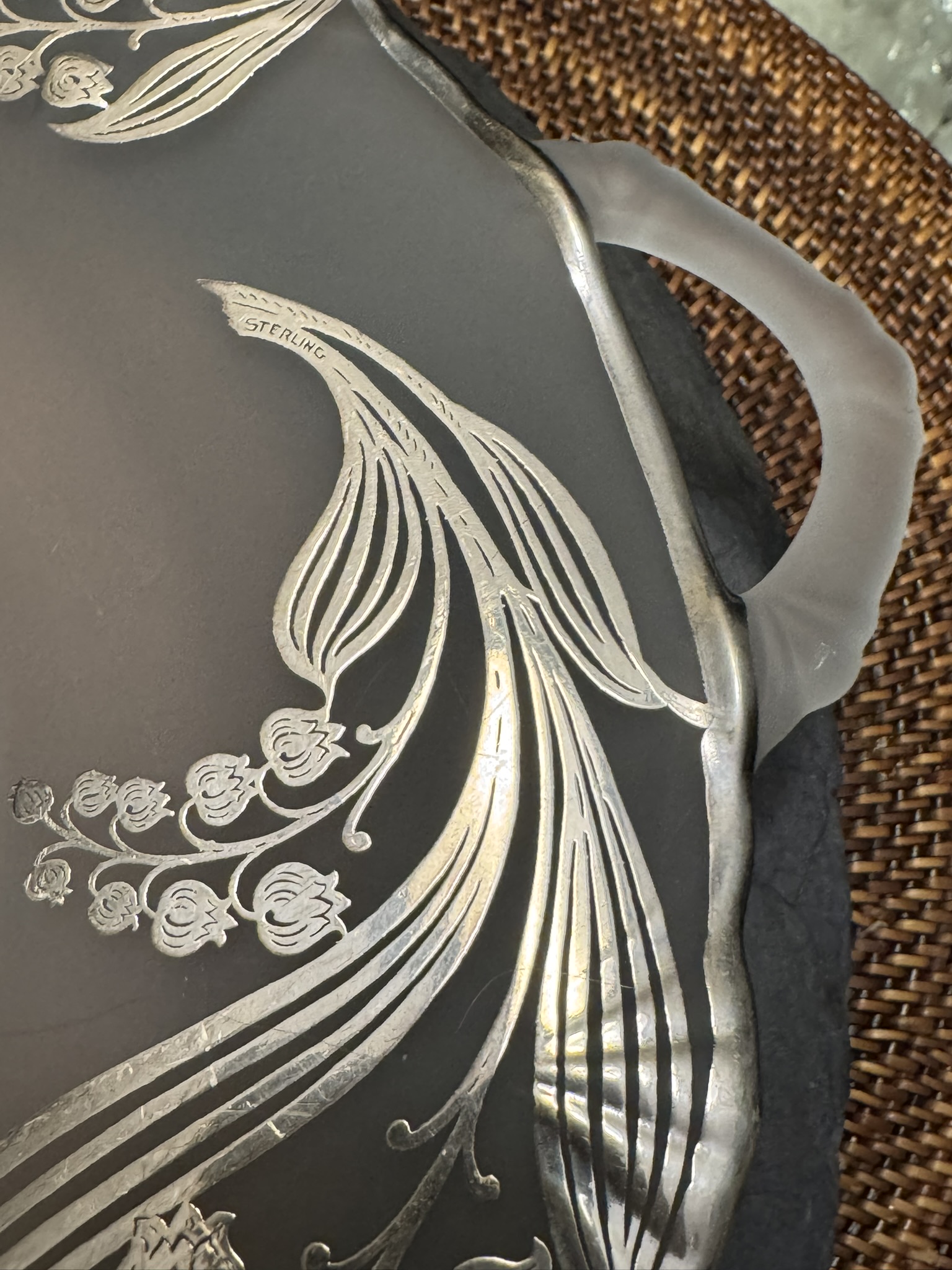 Close-up of a decorative tray handle. The tray features frosted glass with an ornate, etched silver Art Nouveau floral and swirl design. The word “STERLING” is engraved into the silver. The tray sits on a woven brown surface.