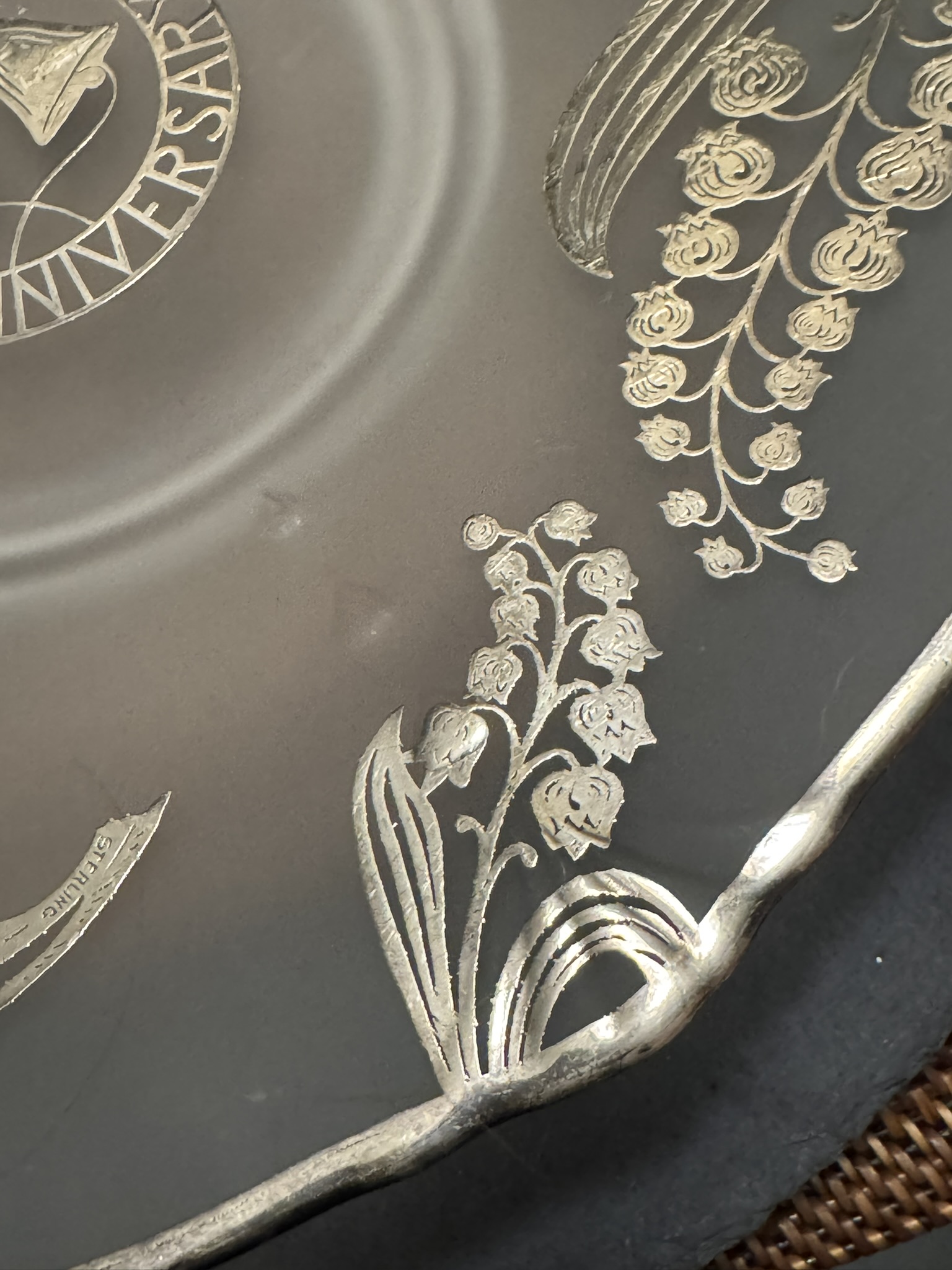 Close-up of an etched glass plate with intricate floral designs, including lily of the valley flowers and leaves. The plate’s edge is scalloped and silver-rimmed. Part of a circular inscription and other decorative engravings are visible in the background.