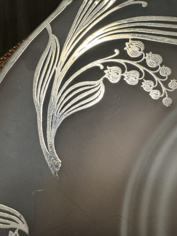 A close-up of a gray plate with a raised, silver etched floral design featuring delicate curved leaves and clusters of small bell-shaped flowers along the rim. The metallic decoration contrasts with the smooth, matte surface of the plate.