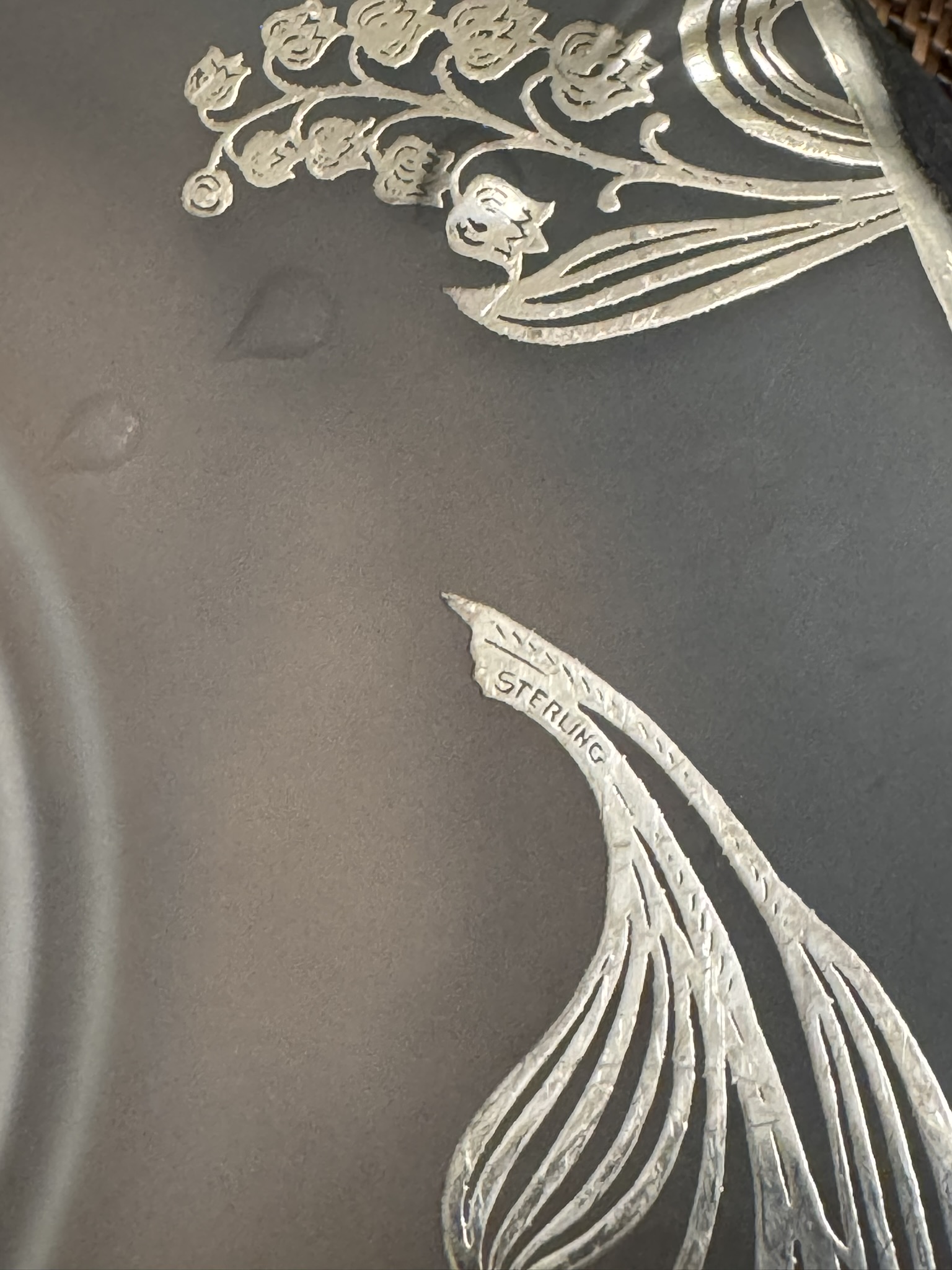Close-up of an engraved floral design on a metallic surface. The etched pattern includes leaves and flowers, with detailed lines. One leaf-like section is inscribed with the word “STERLING,” indicating the item is made of sterling silver.