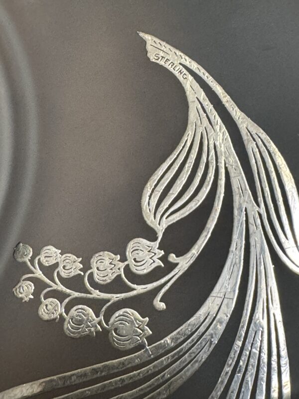 A close-up of a silver etched design on a dark surface features an elegant floral motif and flowing lines. The word STERLING is engraved near the top right, identifying the material as sterling silver.