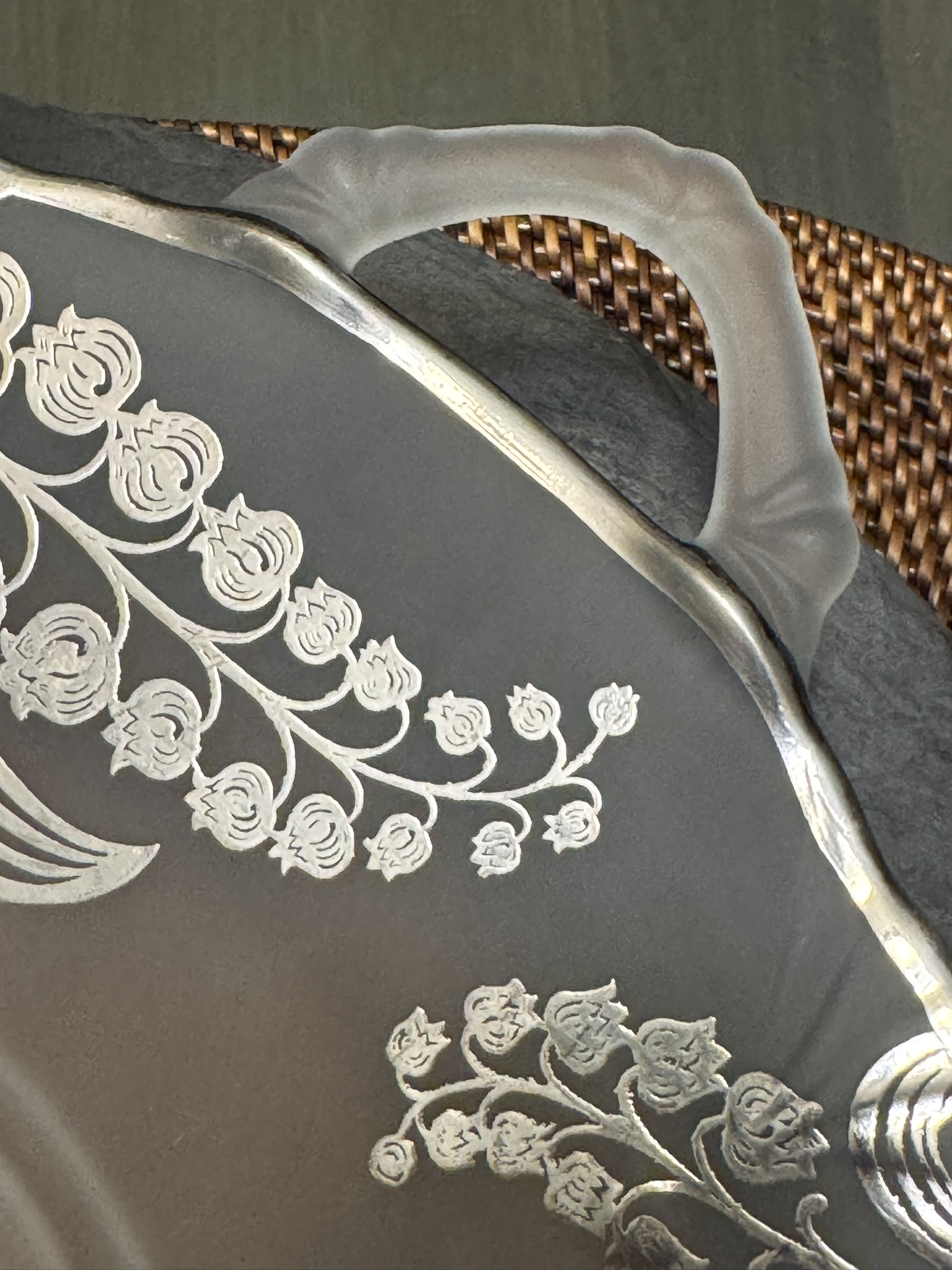 Close-up of a frosted glass plate with an ornate silver floral design. The plate has a curved, frosted handle and rests on a woven brown placemat. The intricate flowers and vines are etched in silver on the glass surface.