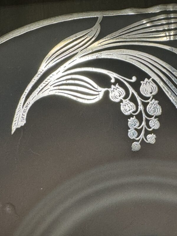 A close-up of a decorative silver plate edge shows an ornate engraved floral design with thin, curved leaves and hanging buds. The word “STERLING” is etched along one of the leaf stems, indicating the silver’s authenticity.