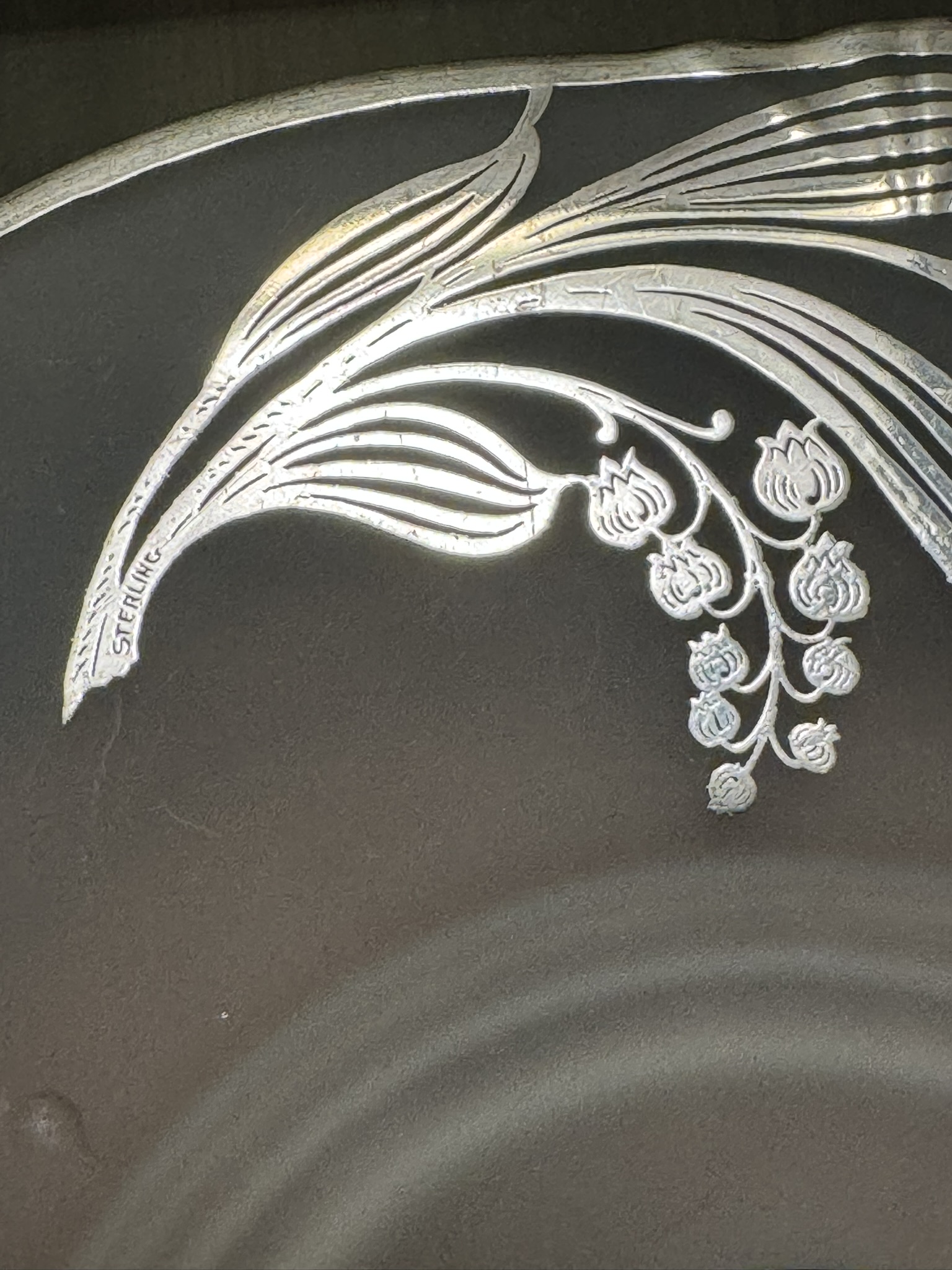 A close-up of a decorative silver plate edge shows an ornate engraved floral design with thin, curved leaves and hanging buds. The word “STERLING” is etched along one of the leaf stems, indicating the silver’s authenticity.