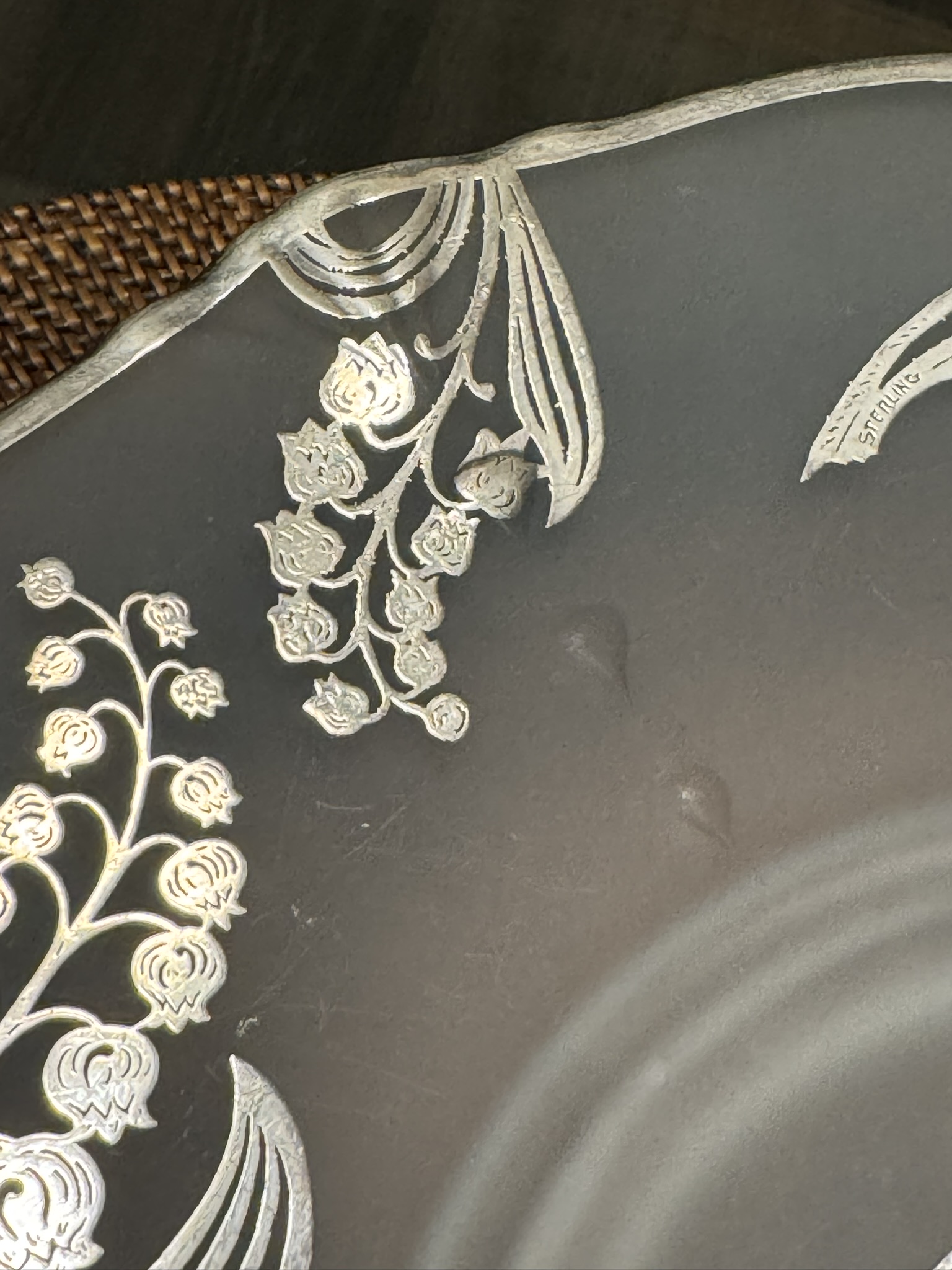 A close-up of a dark, round plate with an ornate silver floral and leaf pattern along the rim. The plate edge is scalloped, and part of a woven brown placemat is visible in the background. The surface shows subtle circular ridges and two small dents.