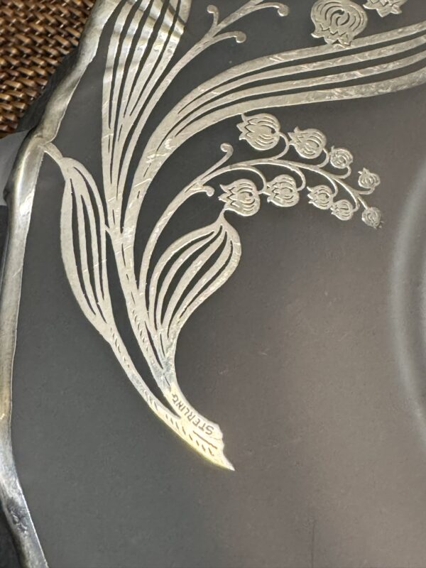 A close-up of a metallic object with an engraved floral design featuring curved leaves and small blooming flowers. The word STERLING is stamped near the base of a leaf, indicating the material is sterling silver. The background is dark and smooth.