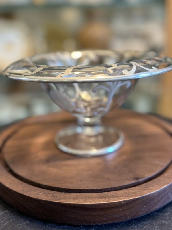A silver pedestal bowl with intricate decorative patterns on the rim sits on a round wooden board. The background is softly blurred, highlighting the ornate details of the bowl and the rich texture of the wood.