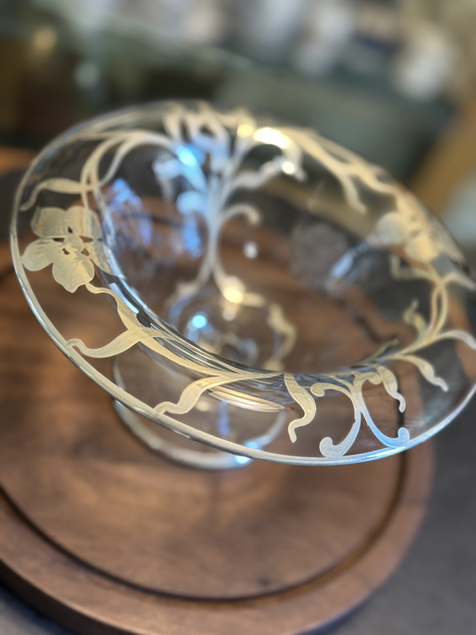 A clear glass bowl with an ornate, frosted floral and vine pattern around the rim sits on a round wooden base. The background is softly blurred, drawing focus to the intricate design of the bowl.