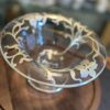 A clear glass pedestal bowl sits on a wooden surface. The bowl has a flared rim and is decorated with delicate, frosted floral and vine patterns around the edge. The background is softly blurred, highlighting the bowl’s elegant design.
