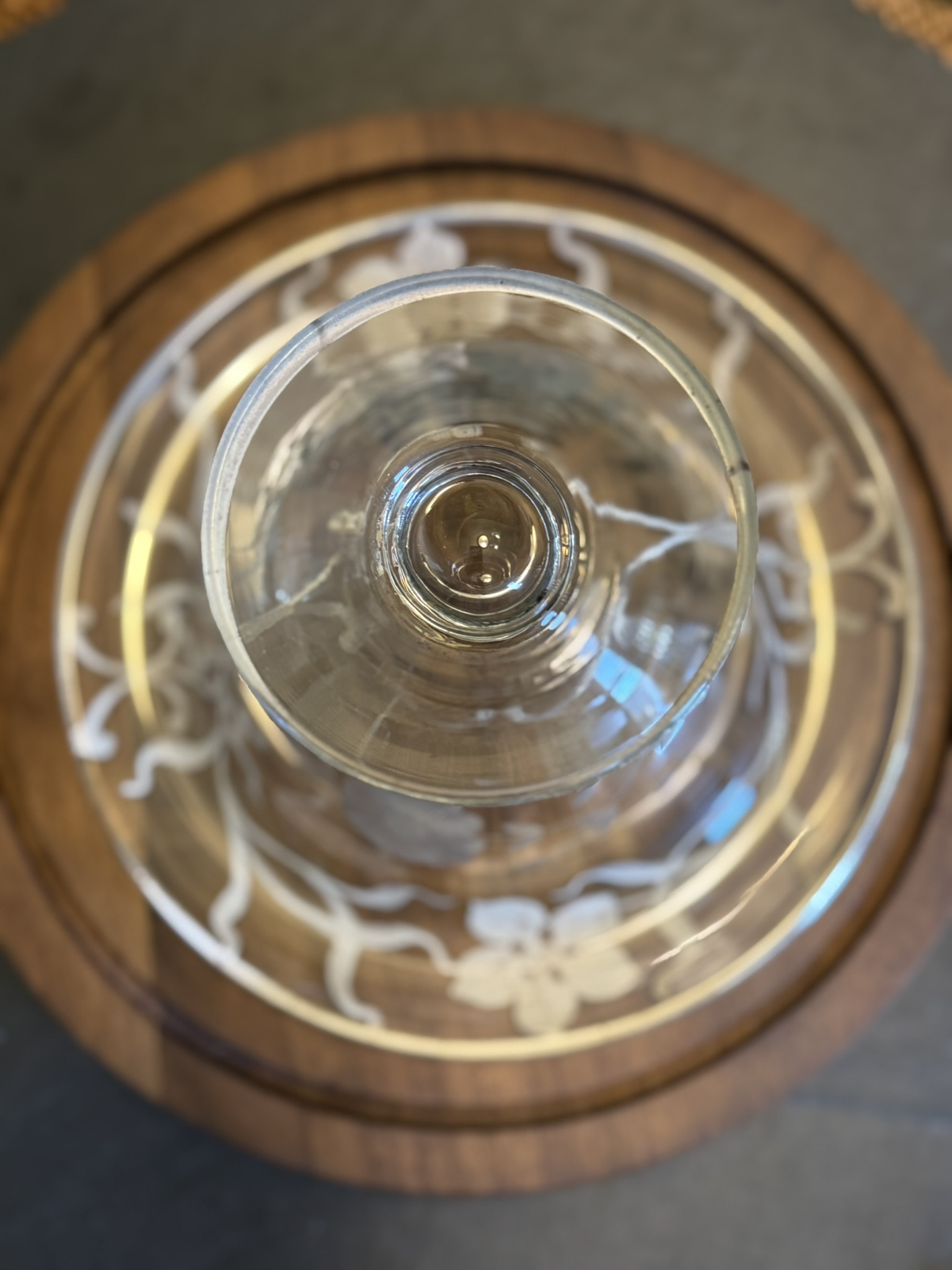 A clear glass viewed from above sits on a wooden tray with a decorative white floral pattern. The glass is centered, creating concentric circular patterns with the tray and floral design. The background is softly blurred.