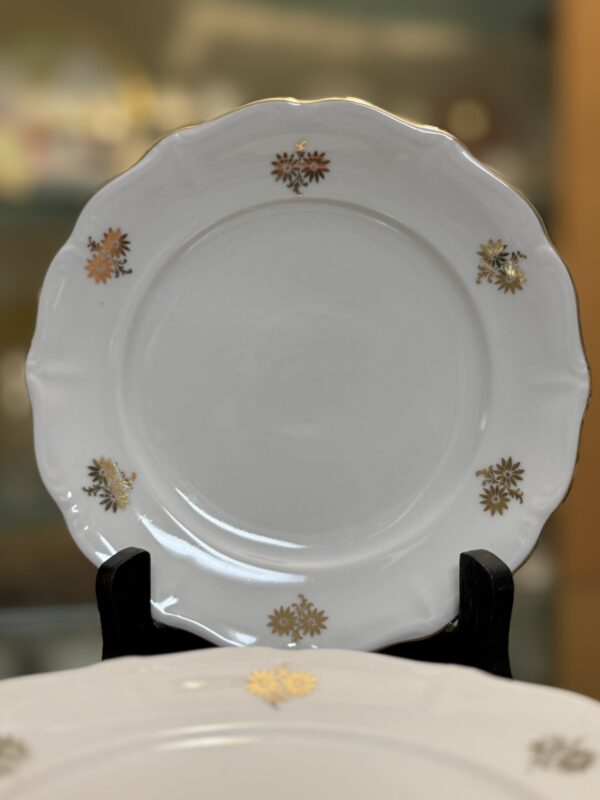 A white porcelain plate with a scalloped edge, decorated with small gold floral motifs evenly spaced around the rim, is displayed upright on a black stand. The background is softly blurred, emphasizing the elegant, glossy plate.