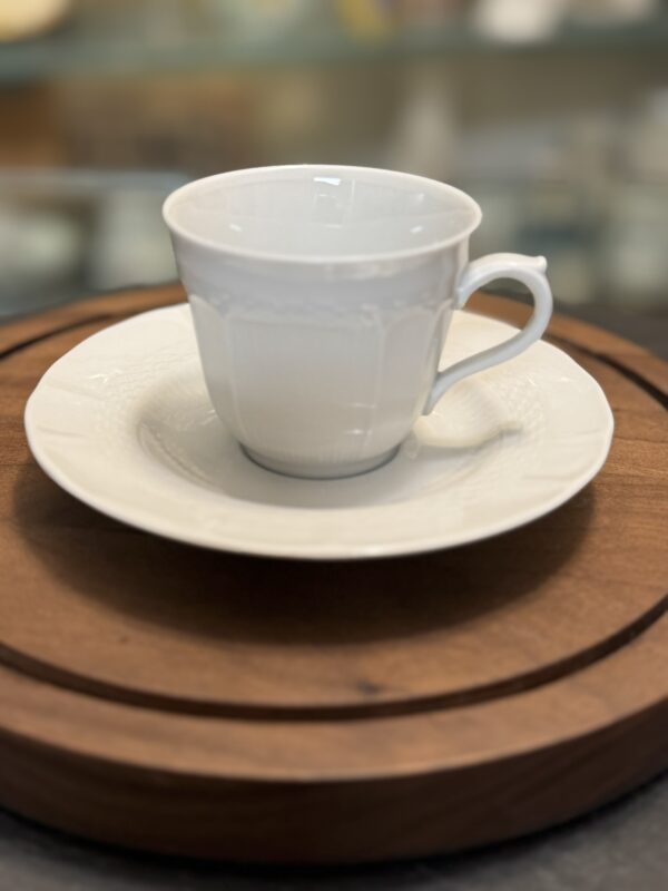A white porcelain teacup sits on a matching saucer, both featuring subtle embossed patterns. The cup and saucer rest on a round wooden tray. The background is softly blurred, focusing attention on the elegant tea set.
