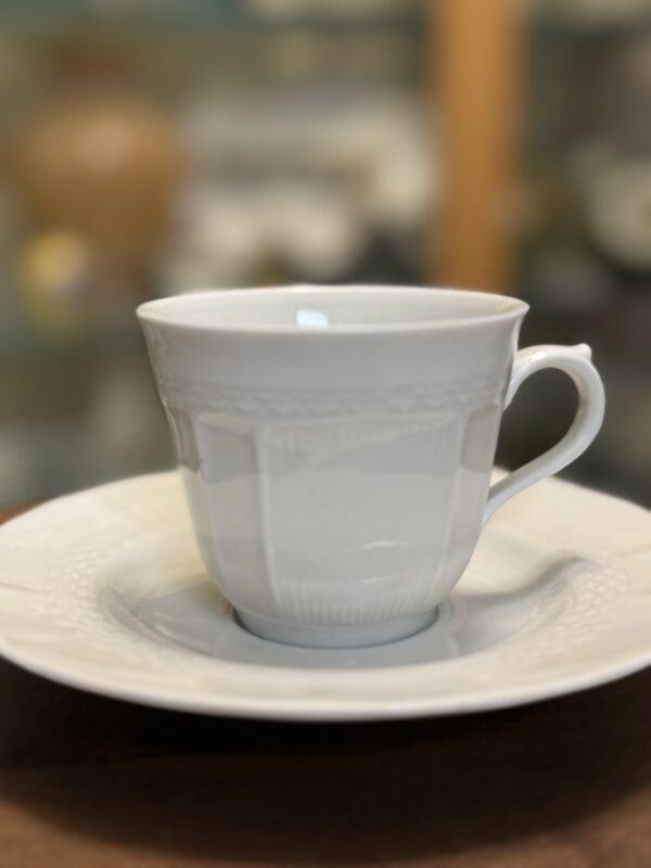 A white porcelain teacup with a decorative textured pattern sits on a matching saucer. The background is softly blurred, highlighting the elegant, classic design of the cup and plate. The handle is delicately curved.