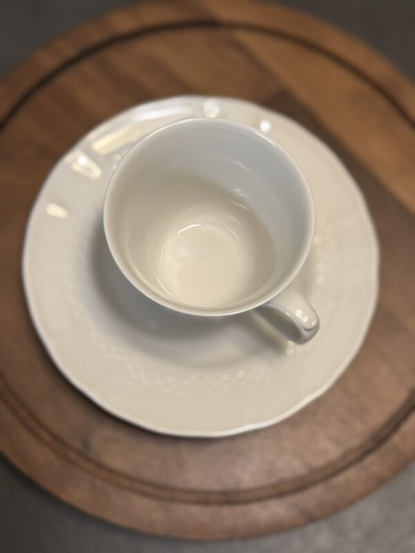 An empty white ceramic teacup sits on a matching white saucer, both with subtle textured patterns. The set is placed on a round wooden surface with two visible circular grooves. The photo is taken from above, centered on the cup.