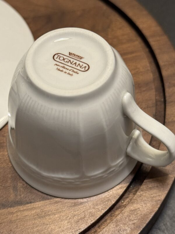 A white ceramic cup placed upside down on a wooden surface. The cup has a curved handle and the base displays the brand name Tognana, along with text stating “porcellana d’Italia” and “Made in Italy.” Part of a white saucer is visible nearby.