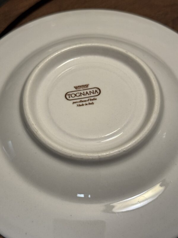 The image shows the underside of a white plate with a circular ridge. At the center, there is a brown logo and text that reads: “Tognana, porcelain d’Italia, Made in Italy.” The surface is smooth and glossy, reflecting light.