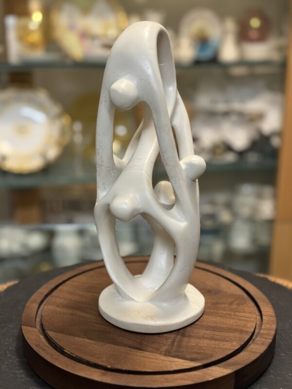 A smooth, abstract white sculpture with intertwined, looping shapes and three rounded nodules stands on a circular wooden base. In the background, glass shelves display decorative porcelain and ceramic plates and objects, softly blurred.