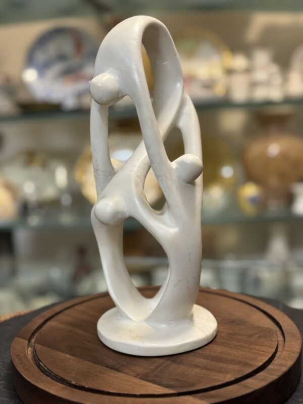 A smooth, white abstract sculpture with flowing, intertwined loops and four spherical shapes is displayed on a round wooden base. In the blurred background, glass shelves hold various decorative plates and objects.