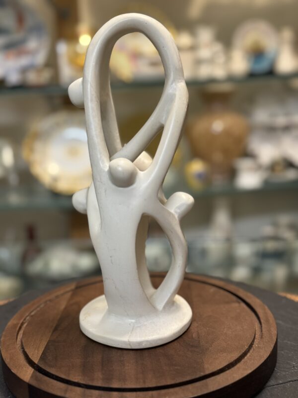 A smooth white abstract sculpture shows three stylized human figures with elongated limbs. They form a circular, interconnected shape, standing on a round base. The background is softly blurred, showing shelves with decorative plates and glass objects.