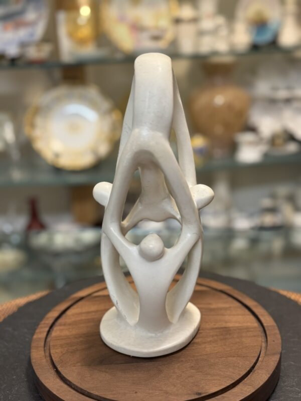 A smooth, white abstract sculpture of three human figures holding hands in a circle, forming an arch. The sculpture sits on a round wooden base. In the blurred background, shelves display various decorative plates and glassware.