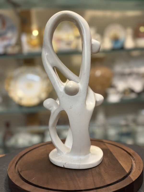 A smooth, abstract white sculpture with intertwined, looping curves and three rounded protrusions sits on a wooden base. The background features blurred shelves displaying decorative plates and collectibles.