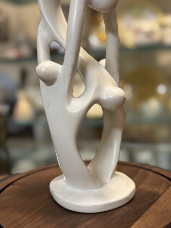 A smooth, abstract white sculpture with intertwined, elongated forms and rounded shapes sits on a circular wooden base. The background is softly blurred, showing shelves with various colorful objects.