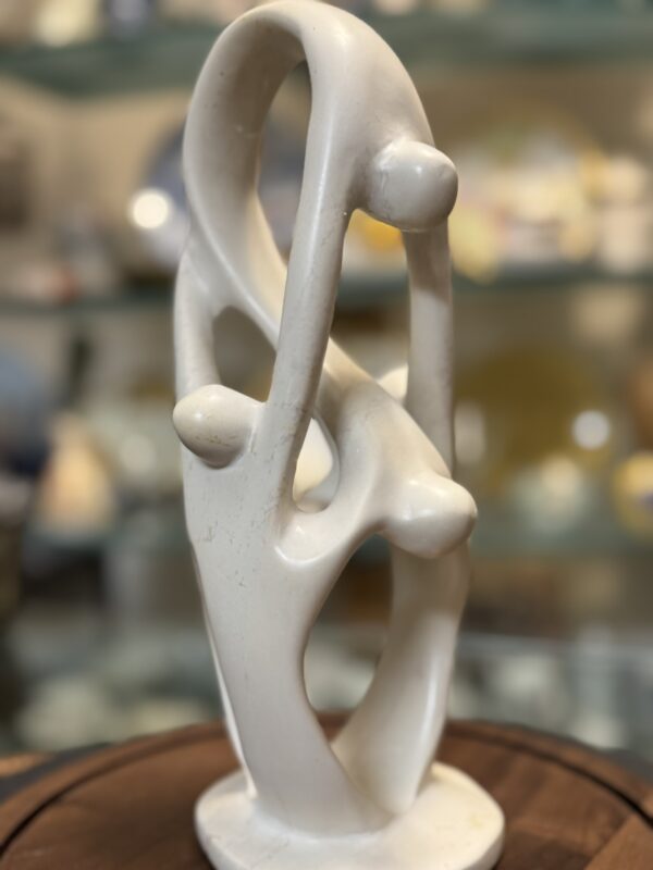 A smooth, abstract white sculpture features three intertwined, elongated forms resembling human figures. The figures are connected and reach toward each other, forming a loop. The background is softly blurred, highlighting the sculpture’s flowing curves.