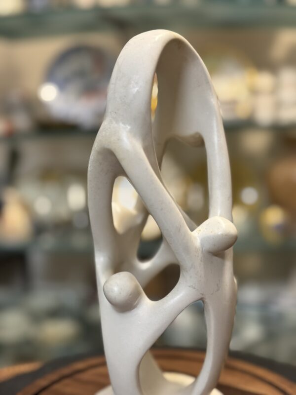 A smooth, abstract white sculpture with oval and circular openings and rounded protrusions stands on a wooden base. The background is softly blurred, showing various ceramics and plates on glass shelves.