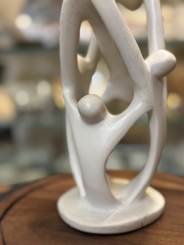 A smooth, white abstract sculpture featuring interconnected human figures holding hands in a circular formation, standing on a small round base. The background is blurred, highlighting the sculpture’s flowing, organic shapes and communal theme.