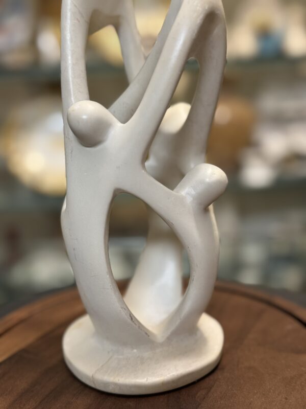 A smooth, cream-colored abstract sculpture of three intertwined human figures with elongated limbs stands on a round wooden surface. The background is softly blurred, showing indistinct shelves with various decorative objects.