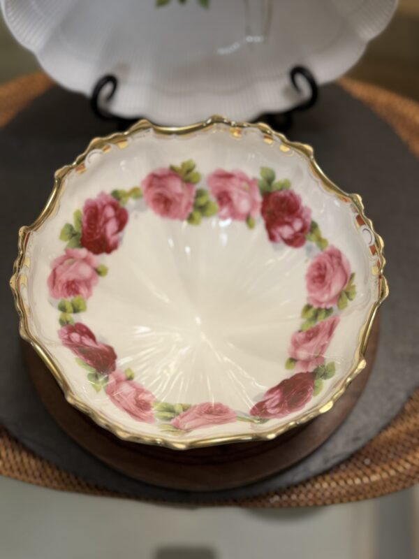 A decorative porcelain bowl with scalloped, gold-rimmed edges, featuring a wreath of pink and red roses with green leaves around the interior. The bowl sits on a round woven mat, with another white platter visible in the background.