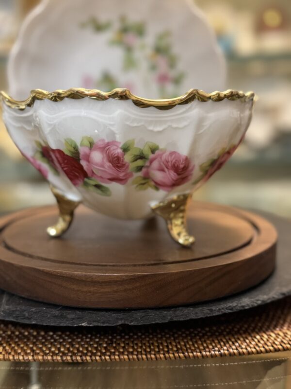 A close-up of an ornate porcelain bowl with gold-trimmed scalloped edges and three gold feet, decorated with pink and red roses and green leaves. The bowl rests on a wooden stand, with another matching dish blurred in the background.