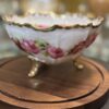 A delicate, floral-patterned porcelain bowl with gold trim and three ornate gold legs sits on a round wooden stand. The bowl features pink roses and green leaves, with a scalloped rim, and is displayed against a softly blurred background.