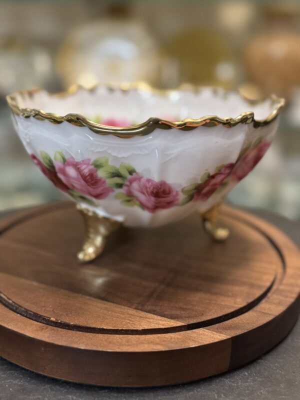 A delicate, floral-patterned porcelain bowl with gold trim and three ornate gold legs sits on a round wooden stand. The bowl features pink roses and green leaves, with a scalloped rim, and is displayed against a softly blurred background.